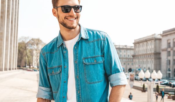 portrait of handsome smiling stylish hipster lumbersexual businessman model. man dressed in jeans jacket clothes. fashion male posing on the street background in sunglasses