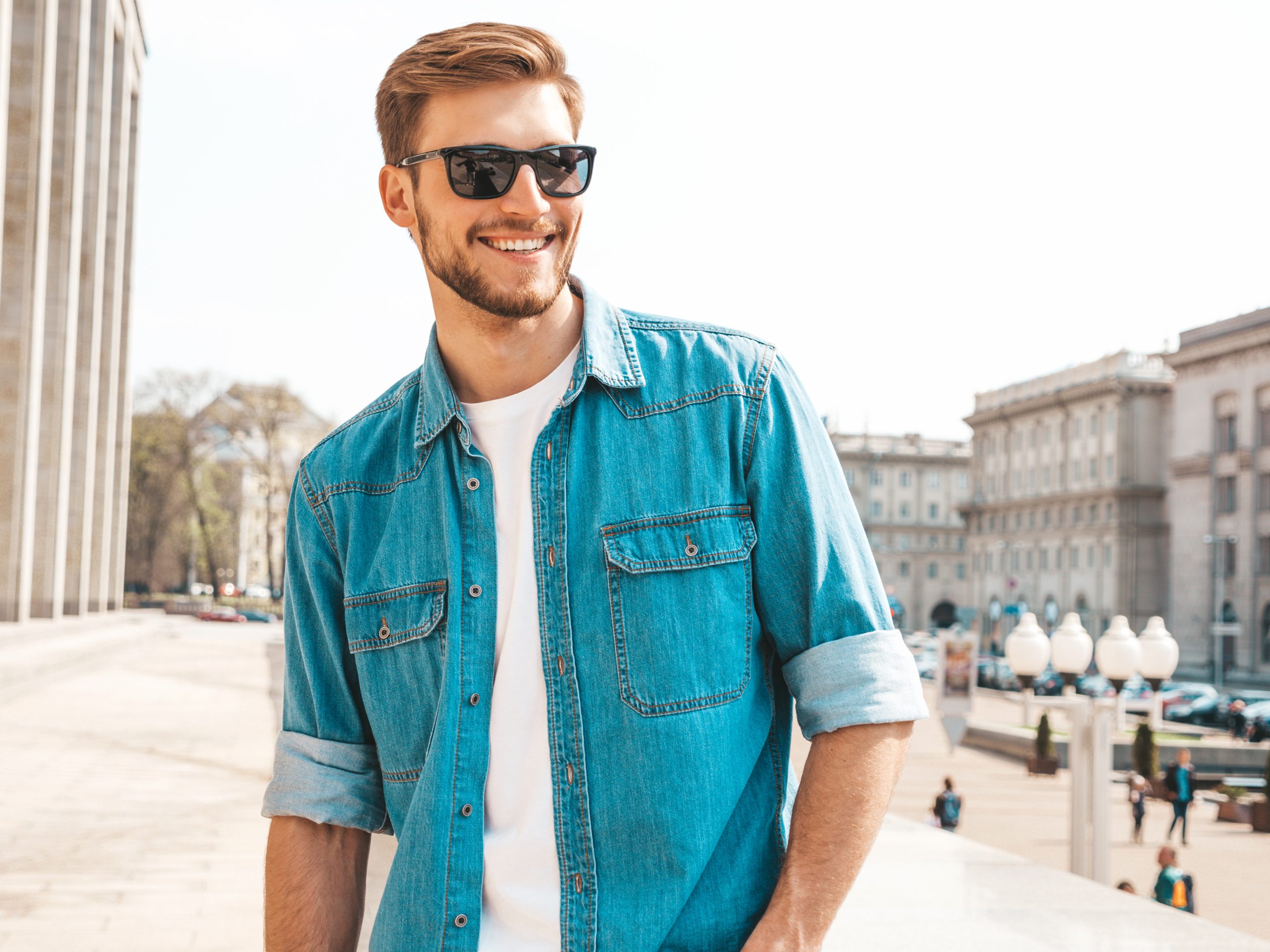 portrait of handsome smiling stylish hipster lumbersexual businessman model. man dressed in jeans jacket clothes. fashion male posing on the street background in sunglasses